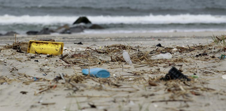 Plastic waste litters sand on a beach, with waves seen in the background.