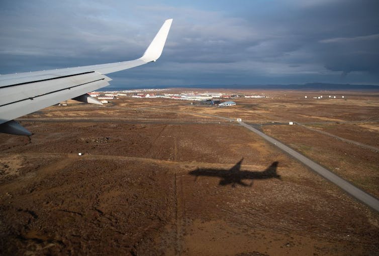 Perspective from a plane window of the plane's shadow against a brown field with the plane's white wing visible on the left side.