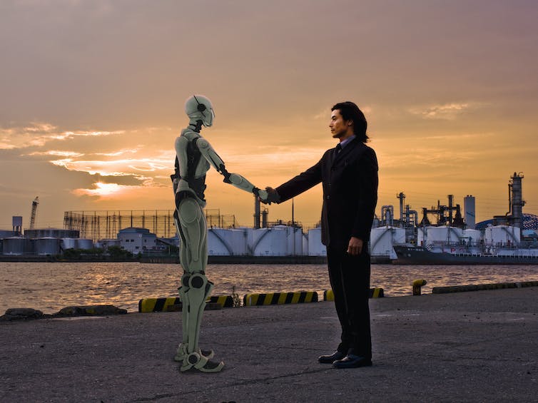 A humanoid robot and a man in a business suit shake hands while standing on an industrial waterfront