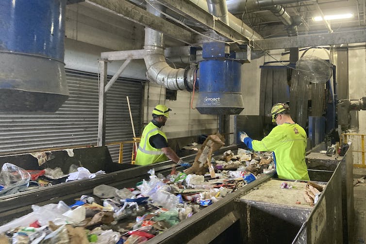 Two workers, in bright yellow, stand at a conveyor belt covered in plastics in a recycling facility.