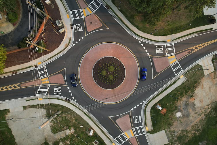 A bird's-eye view of a roundabout, with a pink circular center with grass in the middle, and four roads converging from north, south, east and west.