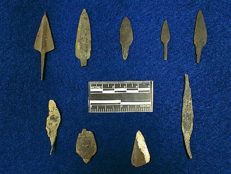 Nine copper projectile points of varying shapes rest on a fabric backdrop, with a scale bar between them.