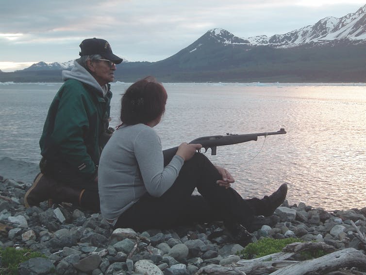 man and woman sit at rocky edge of water, she holds a rifle