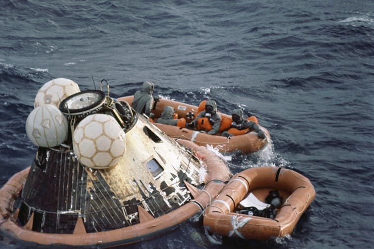 Two small rafts, one full of crew members, float next to a metal capsule.