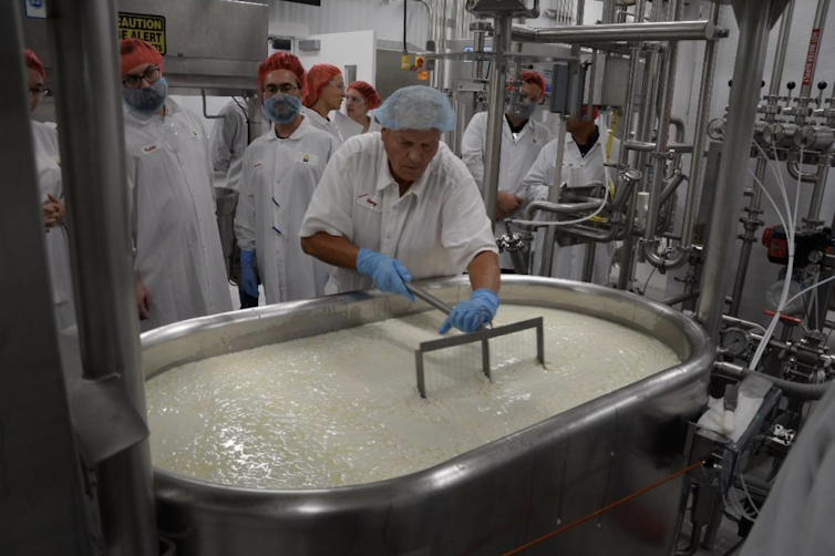 A man in a white lab coat, hairnet and gloves pulls a device through a large tub of white liquid.