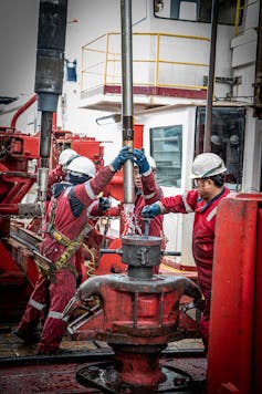 people in protective gear and hard hats on a ship's deck, raising a long metal tube with water coming out the bottom