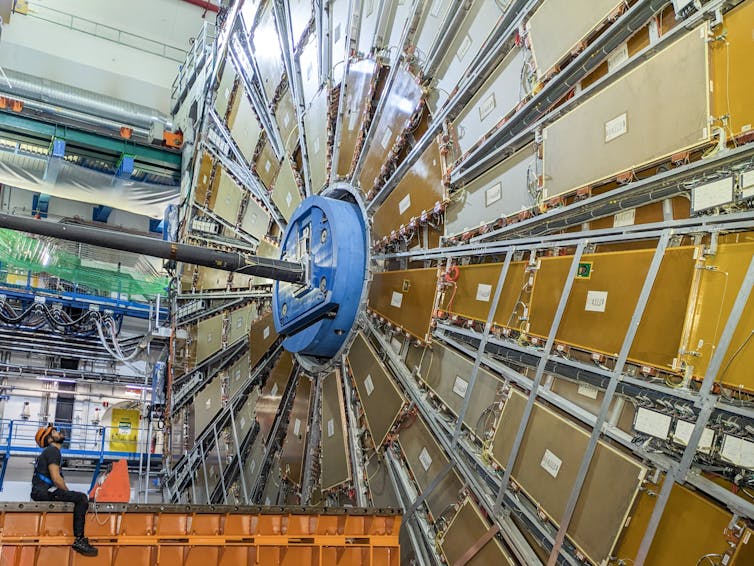 Male technician sitting on scaffolding and facing one side of the ATLAS experiment at CERN