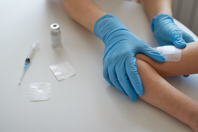 Gloved hands of clinician placing bandaid on child's arm, a syringe and vaccine vial beside them