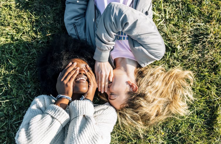 Two smiling people lying on grass, one with hands over eyes and the other whispering into their ear