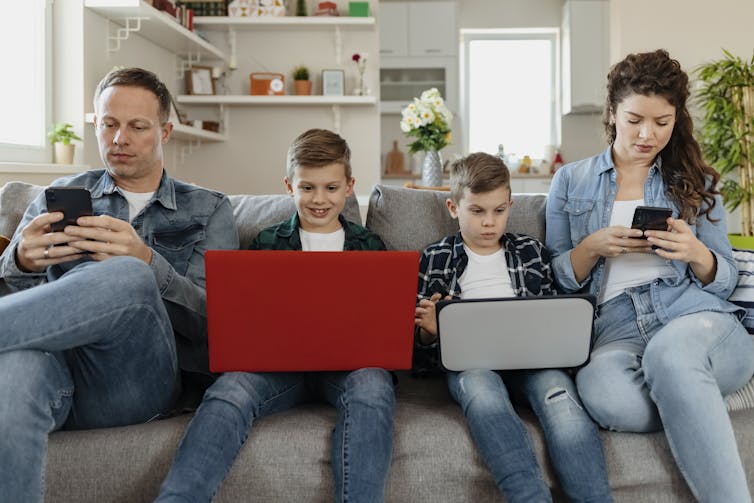 man and woman on smartphones sit on couch with two kids using laptops, all engrossed in screens