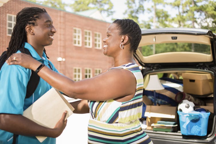 woman with arms on younger man's shoulders by back of a packed car, both smiling