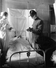 masked nurse and military man stand above patient in bed