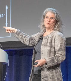 Andrea Ghez in a gray cardigan lecturing in front of a large screen showing a physics diagram.