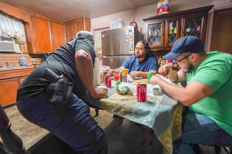 man wearing a holstered gun sitting down to eat at kitchen table with two others