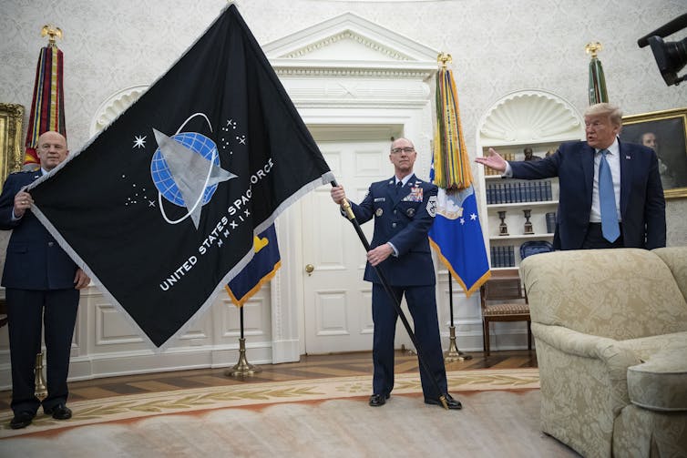 A commander in military uniform waves a black flag with the emblem of the US Space Force (an arrow pointing up in front of a sphere representing the Earth).