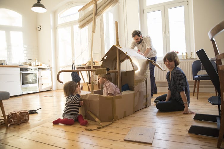 two adults and two small children play with an arrangement of cardboard boxes in a brightly lit room with hard flooring