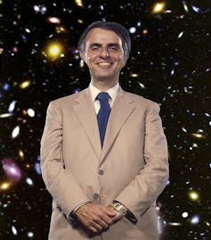 Carl Sagan poses before a backdrop that shows the stars and galaxies of space.