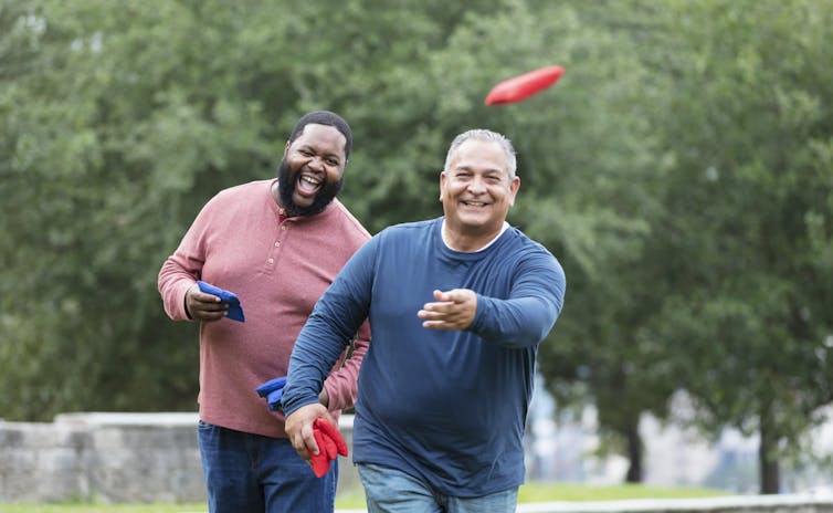 two men in a park grin as one tosses a bean bag