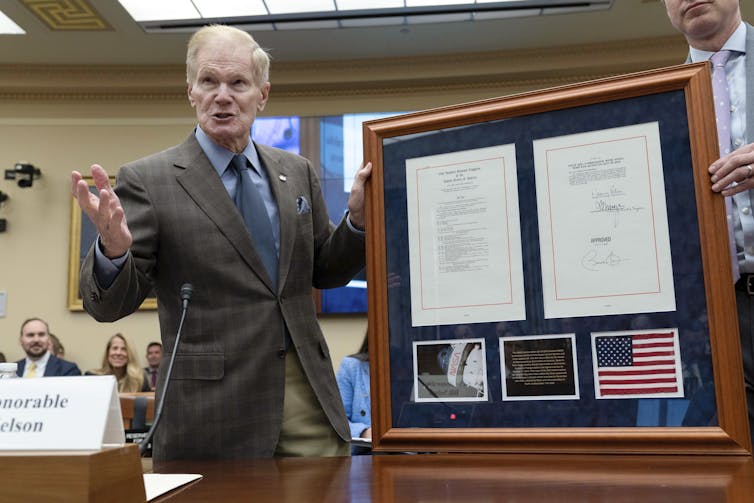 Bill Nelson, wearing a suit jacket and tie, holds a frame containing two documents, a photo of the lunar surface and the American flag.