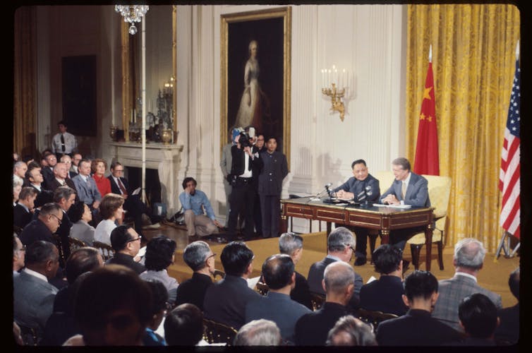 two leaders sit at table in front of Chinese and American flags with press corps in front of them