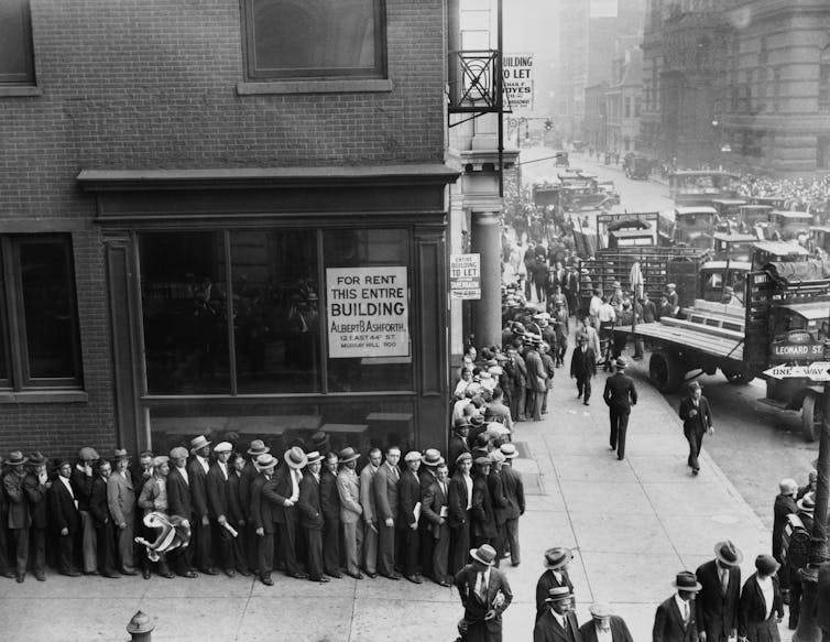 black and white picture of dozens of men in suits and hats lined up on a city street corner