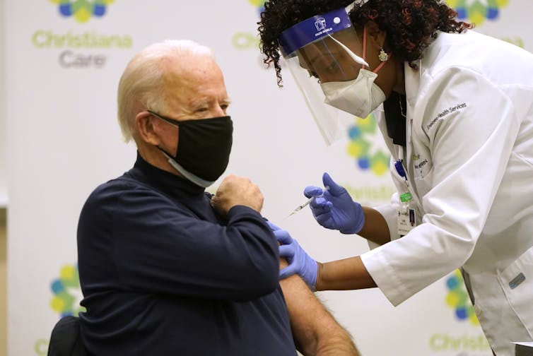 Seated and masked Biden gets a shot in his arm from a masked medical worker