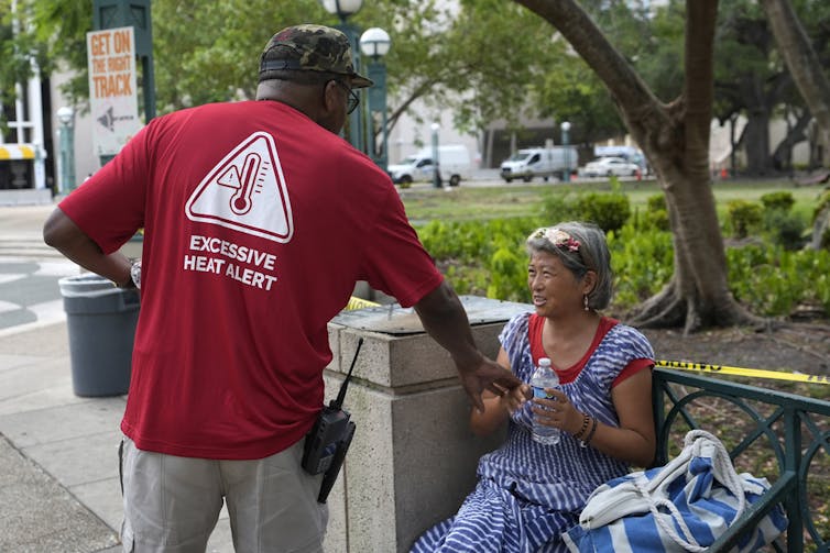 Person wearing a shirt reading 'EXCESSIVE HEAT ALERT' handing water bottle to older adult sitting outside