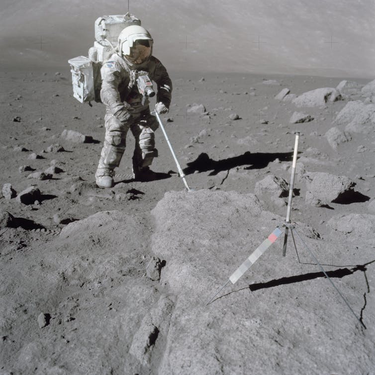 An astronaut standing on the lunar surface and using a scoop to retrieve samples.