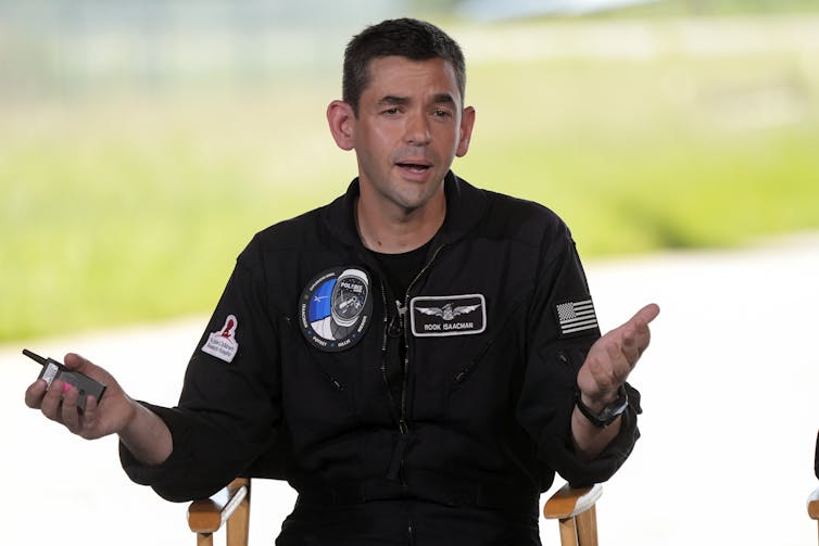 A man with short dark hair wearing a black jumpsuit with an American flag patch and the Polaris mission insignia.