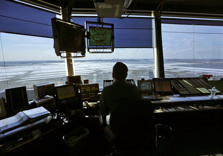 a man sits in silhouette at a panel of computer screens as he looks out of the large windows onto a runway