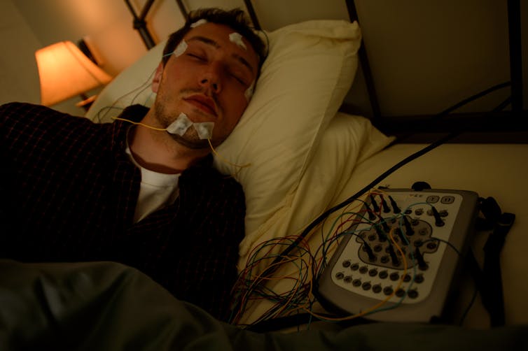 man lying in bed with little electrodes stuck around his face and head, wires run to a machine