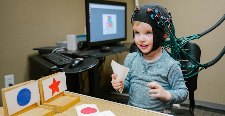 Child wearing hat with sensors on it next to cards with shapes of different colors