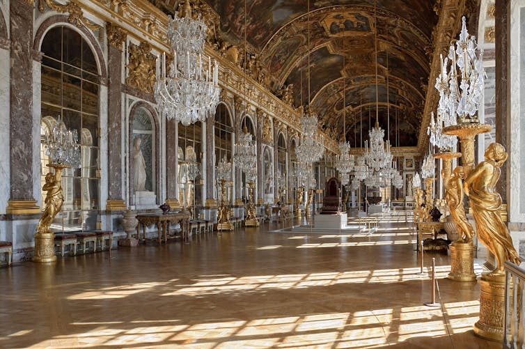 A large hall lined with mirrors, with a painted ceiling, statutes and large chandeliers.
