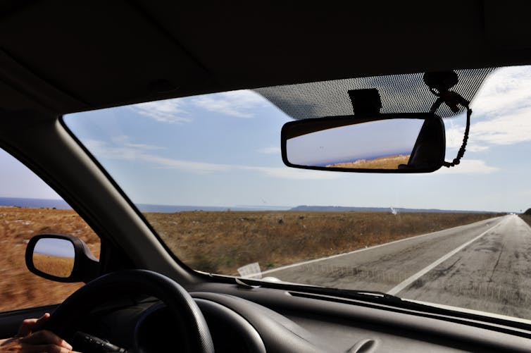 The view from the driver's seat of a car, showing the top of a steering wheel, the windshield view and the rearview mirror.