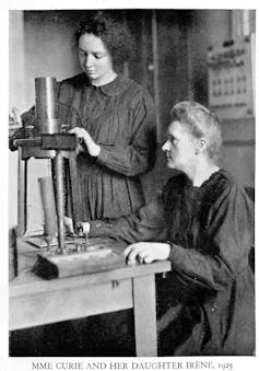 Marie Curie sits at a table with scientific equipment on it. Irene Curie stands next to her, fiddling with the equipment.