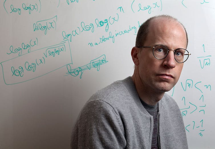 A bald man wearing glasses and a gray top poses solemnly in front of a whiteboard with equations in green marker.
